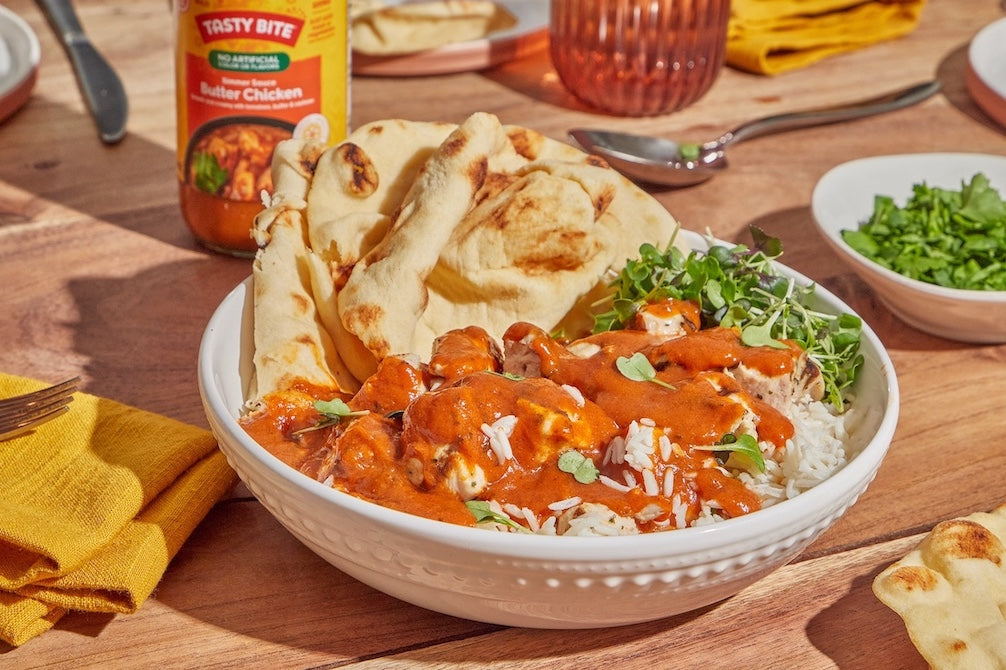 Bowl of rice with Tasty Bite Butter Chicken Simmer Sauce, served with naan and greens on a wooden table.