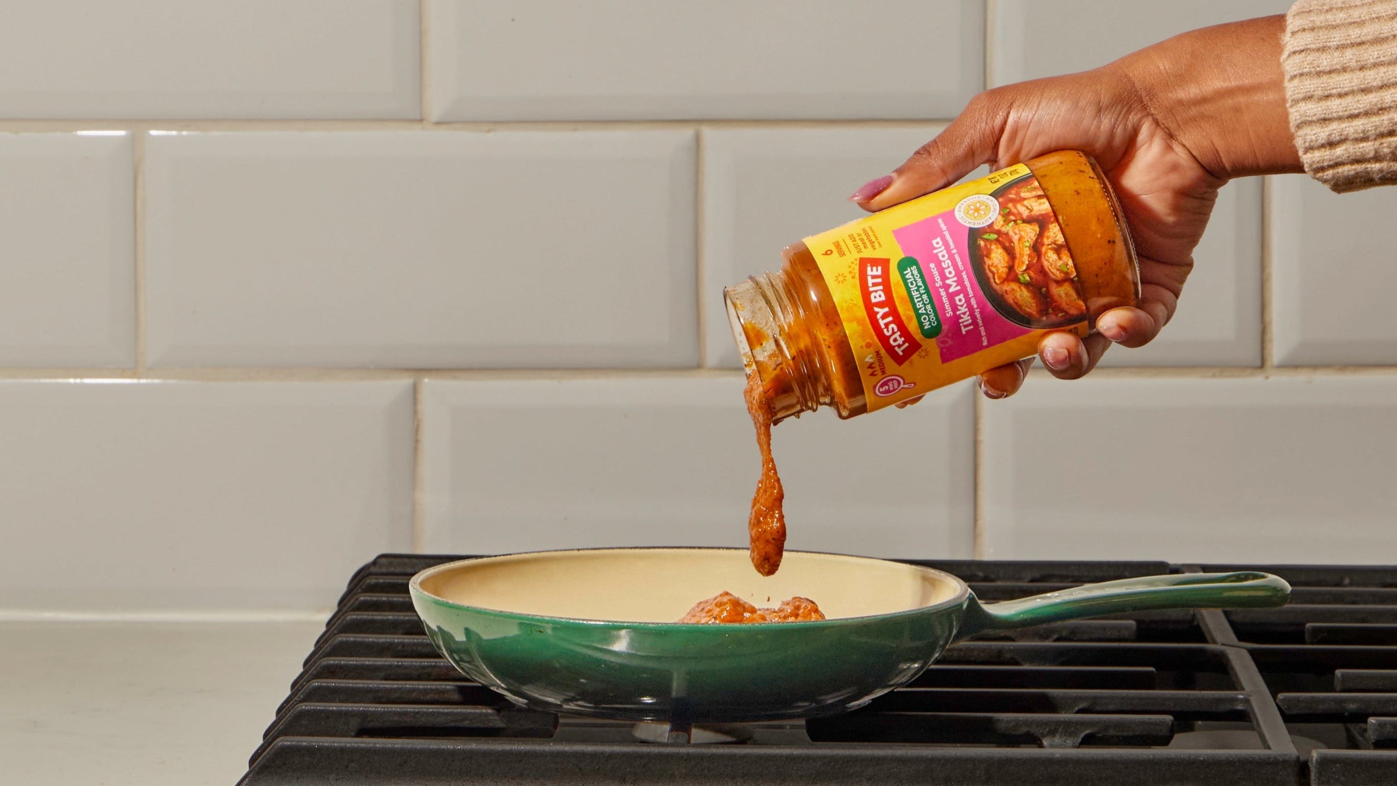 Hand pouring Tasty Bite Tikka Masala Simmer Sauce into a skillet on a stovetop. Indian cooking sauce ready-to-use for quick homemade meals.